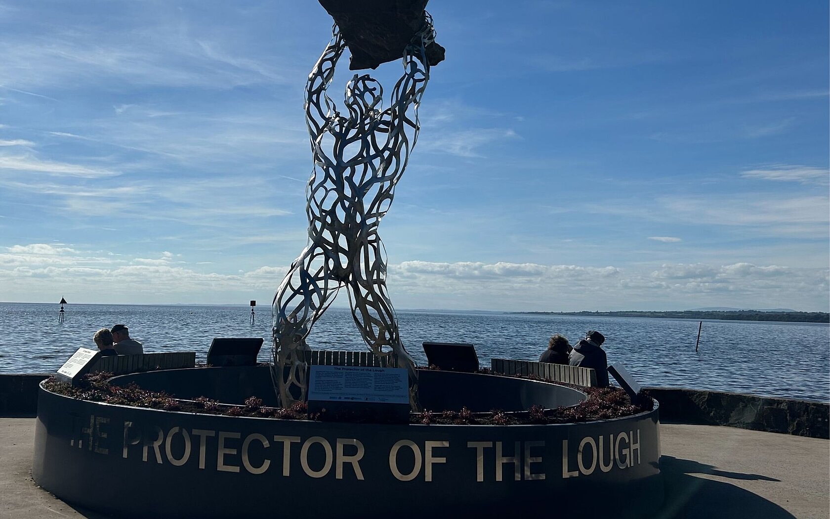 Photo of a statue of the 'protector of Loch Neagh', a humanoid figure holding an outline of the Loch, with the Loch in the background.
